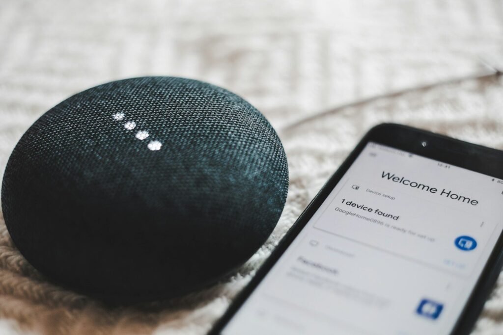 A charcoal Google Home Mini smart speaker next to a smartphone showing a smart home app